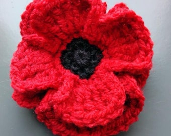May include: A red crocheted poppy flower with a black center. The flower is made with yarn and has a textured surface.