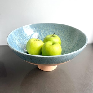K&ouml;nnte beinhalten: Eine blaue Keramikschale mit gesprenkelter Oberfl&auml;che, die auf einem kleinen, hellfarbigen Sockel ruht. Die Schale enth&auml;lt drei gr&uuml;ne &Auml;pfel. Die Schale hat ein breites, flaches Design, das sich zum Pr&auml;sentieren von Obst oder anderen Gegenst&auml;nden eignet.