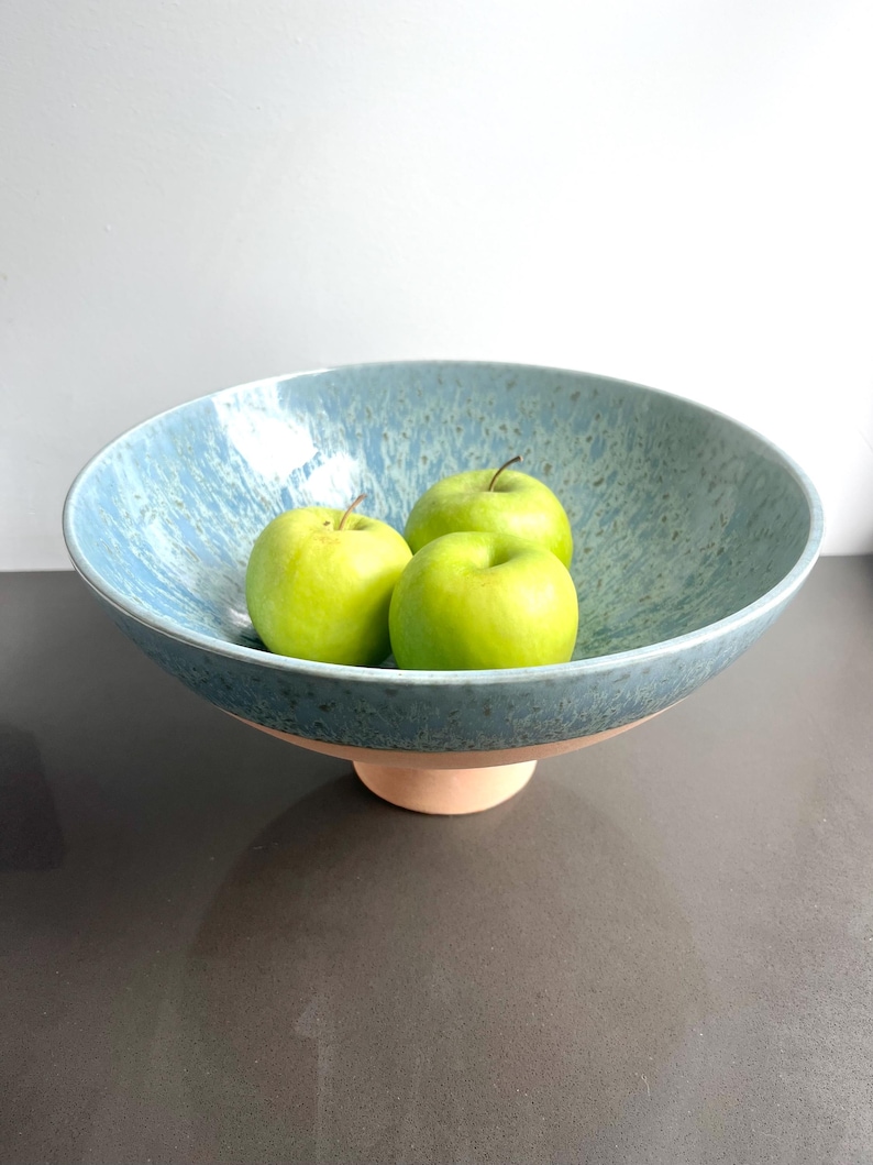 K&ouml;nnte beinhalten: Eine blaue Keramikschale mit gesprenkelter Oberfl&auml;che, die auf einem kleinen, hellfarbigen Sockel ruht. Die Schale enth&auml;lt drei gr&uuml;ne &Auml;pfel. Die Schale hat ein breites, flaches Design, das sich zum Pr&auml;sentieren von Obst oder anderen Gegenst&auml;nden eignet.