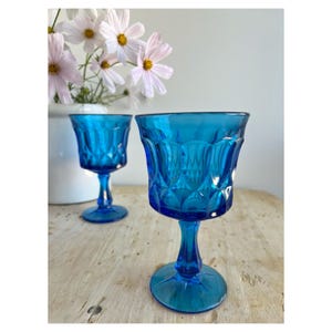 Op de afbeelding: Twee vintage blauwe wijnglazen met diamant- en verticale patroondetails. De glazen staan op een licht houten oppervlak, met een witte vaas met roze en witte bloemen op de achtergrond. De glazen hebben een steel en een ronde voet.