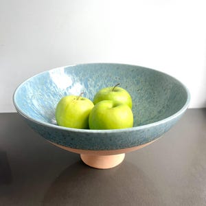 K&ouml;nnte beinhalten: Eine gro&szlig;e, blaue Keramik-Obstschale mit gesprenkelter Oberfl&auml;che, die auf einem hellbraunen Sockel steht. Die Schale enth&auml;lt drei gr&uuml;ne &Auml;pfel. Die Schale ist ein dekoratives St&uuml;ck f&uuml;r die K&uuml;che oder das Esszimmer.