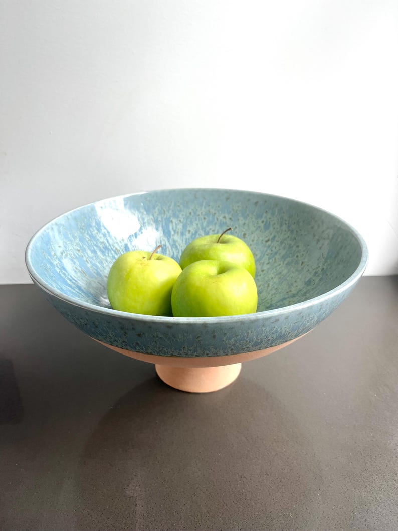 K&ouml;nnte beinhalten: Eine gro&szlig;e, blaue Keramik-Obstschale mit gesprenkelter Oberfl&auml;che, die auf einem hellbraunen Sockel steht. Die Schale enth&auml;lt drei gr&uuml;ne &Auml;pfel. Die Schale ist ein dekoratives St&uuml;ck f&uuml;r die K&uuml;che oder das Esszimmer.