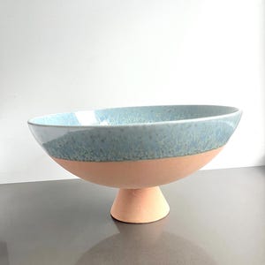 K&ouml;nnte beinhalten: Eine Keramikschale mit hellblau glasierter Kante und pfirsichfarbenem Sockel. Die Schale steht auf einem kurzen, abgerundeten Sockel. Das Innere der Schale ist nicht sichtbar.