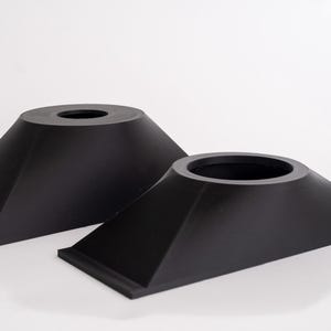 May include: Two black, geometric, cone-shaped objects with a flat base and a circular opening at the top. The objects are made of a matte material and are set against a white background.