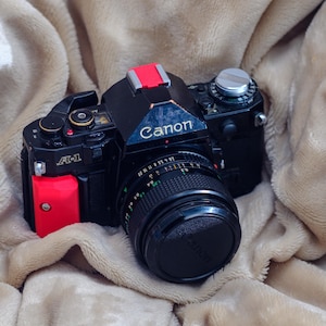 May include: A black Canon A-1 film camera with a lens cap, red accents, and the Canon logo. The camera is on a beige, textured surface.