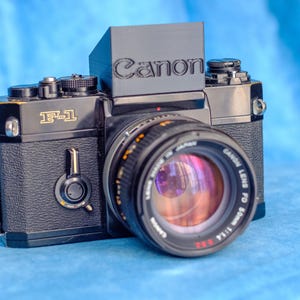 Puede incluir: Una cámara de película Canon F-1 negra con un objetivo Canon. La cámara tiene una cubierta de imitación de cuero negro y una inscripción plateada que dice "F-1". El objetivo es un Canon FD 50mm f/1.8.