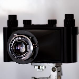 Könnte beinhalten: Eine schwarze Vintage-Kamera mit einem markanten Graphex-Objektiv und silbernen Metalldetails. Der Objektivtubus zeigt den Schriftzug Graphex, Graflex sowie verschiedene technische Spezifikationen. Das Kameragehäuse hat eine rechteckige Form.