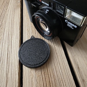 May include: Black Canon ML AF35ML camera with a lens cap on a wooden surface. The camera has the words "Canon ML" and "AF35ML" printed on it. The lens cap is round and textured.