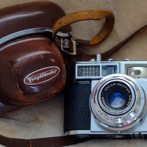 May include: A vintage camera with a brown leather case. The camera is silver and black with a lens that reads "COLOR-SKOPAR". The case has the brand name "Voigtländer" in white lettering. A brown leather strap is attached.