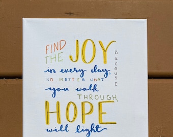 May include: A white canvas with a hand-painted inspirational quote in colorful lettering. The quote reads "Find the joy in every day, no matter what you walk through, hope will light your way. -cb"