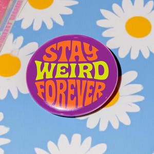 以下が含まれることがあります： 白いヒナギクと黄色の中心部が特徴の、水色の表紙の本。 「STAY WEIRD FOREVER」というテキストが入った紫色のボタンが本の上にあります。 本はピンク色のサテンの表面に置かれています。