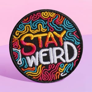 Może przedstawiać: Czarna naszywka haftowana z kolorowymi wirami i tekstem "Stay Weird" w białym, pomarańczowym i czerwonym kolorze.