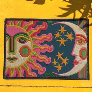 Puede incluir: Parche bordado con un diseño de sol y luna con estrellas. El sol tiene rayos rosas y amarillos, y la luna tiene una cara. El fondo es verde azulado y el parche tiene un borde negro. El parche es rectangular.