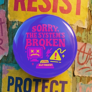 May include: A purple button with the text "SORRY, THE SYSTEM'S BROKEN" and a computer graphic is affixed to a weathered wooden pole covered in colorful posters. The posters read "RESIST" and "PROTECT TRANS YOUTH".