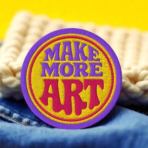 May include: A round patch with the words "MAKE MORE ART" in yellow and pink, surrounded by a purple border. The patch is on blue denim, with a cream-colored knitted item in the background, set against a yellow backdrop.