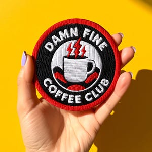 Op de afbeelding: Een ronde patch met een rode rand en de woorden "Damn Fine Coffee Club" in het wit. De patch heeft een witte koffiekop met rode bliksemschichten erboven, tegen een zwarte achtergrond. De patch wordt tegen een gele achtergrond gehouden.