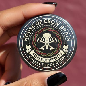 May include: A round, black button with the text "HOUSE OF CROW BRAIN" and "COLLECTOR OF DOOM." The button features a skull and key design with red and green accents. The button is held between fingers against a maroon background.