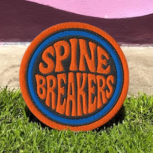 Könnte beinhalten: Ein runder, orangefarbener Aufnäher mit dem Text "SPINE BREAKERS" in orangefarbenen, stilisierten Buchstaben. Der Text befindet sich in einem blauen Kreis, der sich in einem schwarzen Kreis befindet. Der Aufnäher liegt auf grünem Gras, mit einem bunten Wandbild im Hintergrund.