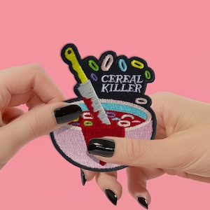 May include: Embroidered patch featuring a bowl of cereal with a knife plunged into it. The patch has the words "CEREAL KILLER" embroidered in white. The cereal pieces are colorful, and the bowl's contents appear to be red.