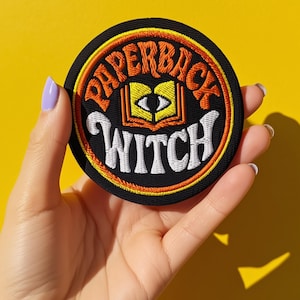 May include: Round black patch with the words "PAPERBACK WITCH" in orange and white embroidery. The patch features an open book with an eye in the center. The background is a bright yellow.