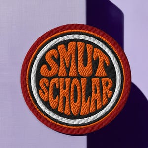 May include: Embroidered patch with the words "SMUT SCHOLAR" in orange, retro-style lettering. The patch is circular, with a black center, a white border, and a red outer edge. The patch is set against a purple background.