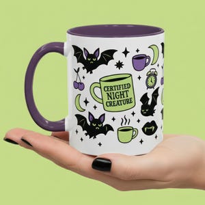 Pu&ograve; includere: Tazza in ceramica bianca con interno e manico viola, con un motivo ripetuto di pipistrelli, lune, stelle e tazze di caff&egrave;. La tazza presenta il testo "CERTIFIED NIGHT CREATURE" su un disegno di tazza di caff&egrave; verde.