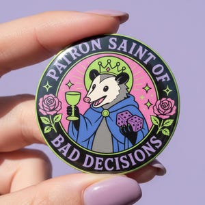 Puede incluir: Un pin redondo de esmalte con un dibujo de un opossum con una túnica azul y una corona, sosteniendo una copa de vino y dados. El pin tiene el texto "PATRON SAINT OF BAD DECISIONS" en un diseño circular, con rosas rosas y detalles verdes.