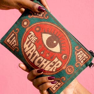 Puede incluir: Una bolsa rectangular verde y roja con el texto "THE WATCHER" y un gráfico de ojo. La bolsa también tiene las palabras "LIFE" y "FATE". La bolsa se sostiene contra un fondo rosa.