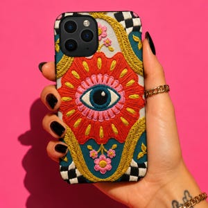 Könnte beinhalten: Dekorative Handyhülle mit gesticktem Design. Die Hülle hat eine rote Blume mit blauem Auge in der Mitte, umgeben von goldenen und türkisfarbenen Akzenten. Die oberen Ecken haben ein schwarz-weißes Schachbrettmuster.