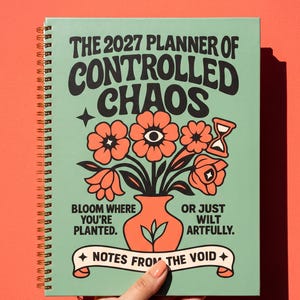 May include: A spiral-bound 2027 planner with a light green cover featuring a central orange vase of flowers, an hourglass, and black text that reads: THE 2027 PLANNER OF CONTROLLED CHAOS, BLOOM WHERE YOU'RE PLANTED, OR JUST WILT ARTFULLY, NOTES FROM THE VOID.