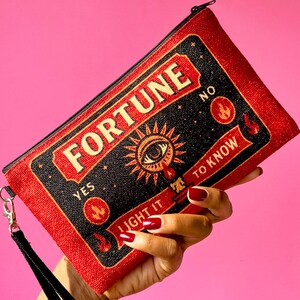 May include: Red and black rectangular clutch with the word "FORTUNE" in gold lettering. The clutch features a zipper closure, a black wrist strap, and the phrases "YES" and "NO" on the top. The design includes an eye and the words "LIGHT IT TO KNOW".