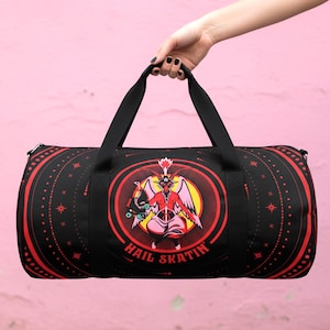 Könnte beinhalten: Schwarze Sporttasche mit roten Akzenten und einer zentralen Grafik einer geflügelten Figur. Die Tasche trägt den Text "HAIL SKATIN" und wird von einer Person gehalten. Die Tasche hat schwarze Griffe und einen Reißverschluss. Der Hintergrund ist eine rosa Wand.
