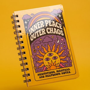 May include: A yellow spiral-bound notebook with the words "INNER PEACE OUTER CHAOS" on the cover, above a sun illustration with three eyes. The design includes a rainbow, clouds, and flowers. The bottom text reads "MEDITATIONS, MANTRAS, AND MELTDOWN NOTES."