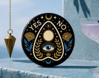 Könnte beinhalten: Ein schwarzes Ouija-Brett mit goldenen und blauen floralen und himmlischen Designs, einschließlich der Wörter "YES" und "NO". Ein goldener Pendel hängt an einer Kette. Ein Cocktail mit Blumengarnitur ist ebenfalls zu sehen.
