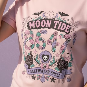 May include: Pink t-shirt with a graphic design featuring the words "MOON TIDE" above the numbers "8647". The design includes bats, moons, shells, stars, roses, and the words "SALTWATER SPELL".