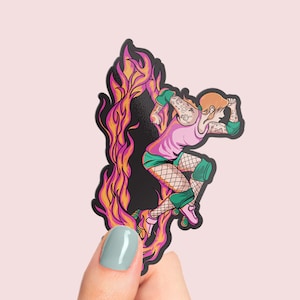 Op de afbeelding: Een sticker met een vrouw die rolschaatst met vlammen achter haar. Ze draagt een roze tanktop, een groene broek en zwarte kousen. Ze heeft tatoeages op haar armen en benen. De sticker is op een zwarte achtergrond.