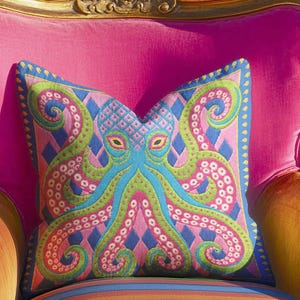 A square decorative throw pillow cover featuring a highly symmetrical bright octopus design. The octopus has a turquoise blue body with a diamond patterned head and eight curled tentacles detailed in bright pink and lime green. The background is a repeating diamond argyle pattern in royal blue and light pink. The artwork is a flat two dimensional print that mimics the textured look of intricate crewelwork embroidery. The pillow features a hidden zipper on the bottom edge. No insert included.