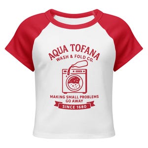 A women's fitted, mid-length micro rib raglan baby tee. The tee's body is solid white, made from a 1x1 micro-rib fabric.  It features short, set-in sleeves and a crewneck collar binding in a bold, contrasting athletic red. Centered on the chest is a vintage-inspired graphic printed in the same red ink. The design is for the "Aqua Tofana Wash & Fold Co." and features an illustration of an old-fashioned washing machine. Text below the machine reads "Making small problems go away" and "Since 1680."