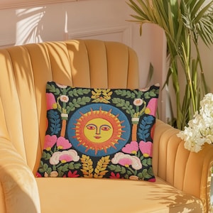 A square folk art sun throw pillow with a dark, charcoal-gray background. The central design is a vibrant celestial sun face in shades of orange, yellow, and red, with defined facial features. The sun is enclosed in a circular blue border with a textured, faux-embroidery pattern. Surrounding the central sun are symmetrical, colorful botanical and floral elements, including pink and white flowers, and green and gold leaves. The overall style is maximalist and bohemian.