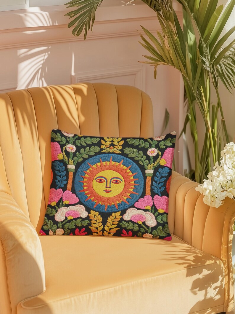 A square folk art sun throw pillow with a dark, charcoal-gray background. The central design is a vibrant celestial sun face in shades of orange, yellow, and red, with defined facial features. The sun is enclosed in a circular blue border with a textured, faux-embroidery pattern. Surrounding the central sun are symmetrical, colorful botanical and floral elements, including pink and white flowers, and green and gold leaves. The overall style is maximalist and bohemian.