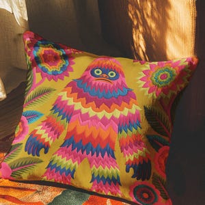 A square faux embroidery pillow featuring a brightly colored maximalist design against a mustard yellow background. In the center stands a stylized, furry Bigfoot or Sasquatch figure rendered in vibrant layers of pink, orange, blue, and lime green with a blue face and yellow eyes. The central creature is surrounded by large, detailed symmetrical pink and blue flowers and leaves. The flat printed fabric creates a highly realistic optical illusion of dimensional crewel embroidery stitches.