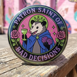 May include: A circular pinback button featuring a cartoon opossum wearing a crown and blue cape, holding a green goblet and purple dice. The design includes pink roses and the text PATRON SAINT OF BAD DECISIONS in a black circular border.