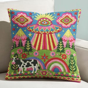 May include: Embroidered square pillow with a whimsical design. The pillow features a blue background with a UFO, cow, rainbow, and floral elements. The pillow has a yellow and pink border.
