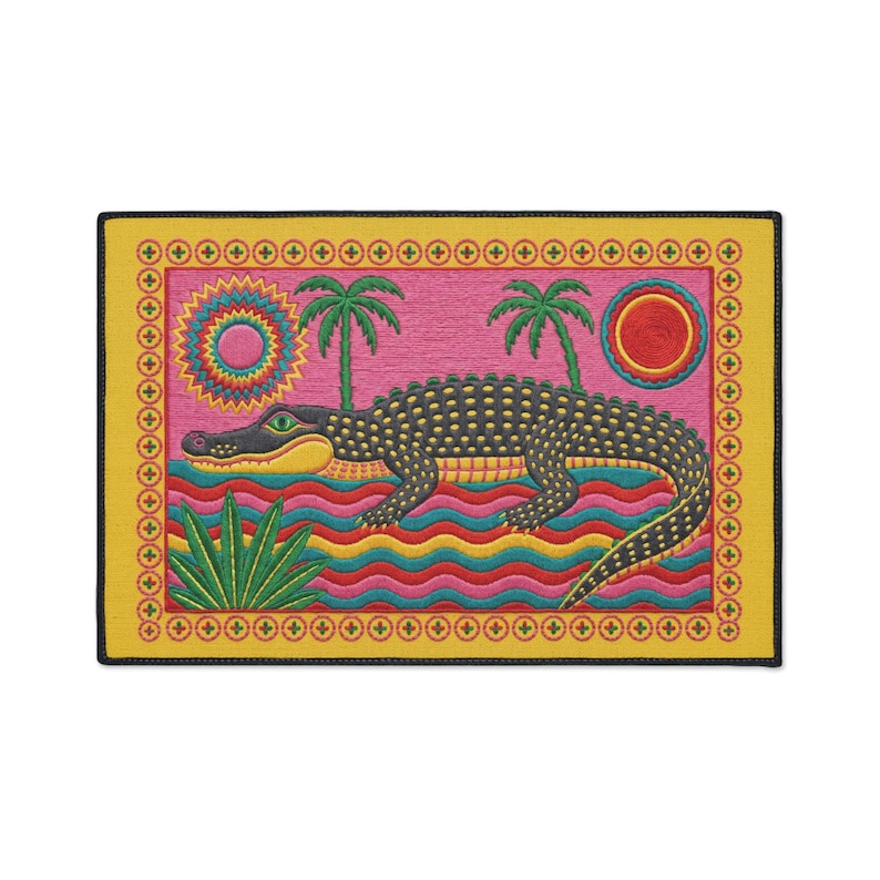 A rectangular doormat featuring a colorful 2D flat print designed to mimic raised, textured yarn embroidery. The central graphic showcases a large dark grey and yellow alligator resting in wavy water stripes of red, bright blue, and yellow. The background is hot pink, decorated with two green palm trees, a leafy green agave plant, and two stylized suns. The entire design is enclosed by a yellow border with repeating circle motifs and finished with a sleek black outer edge border trim.
