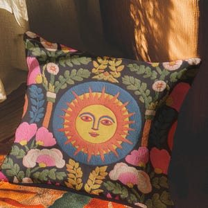 A square folk art sun throw pillow with a dark, charcoal-gray background. The central design is a vibrant celestial sun face in shades of orange, yellow, and red, with defined facial features. The sun is enclosed in a circular blue border with a textured, faux-embroidery pattern. Surrounding the central sun are symmetrical, colorful botanical and floral elements, including pink and white flowers, and green and gold leaves. The overall style is maximalist and bohemian.