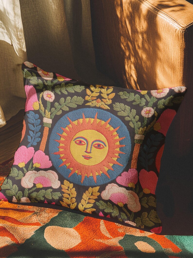 A square folk art sun throw pillow with a dark, charcoal-gray background. The central design is a vibrant celestial sun face in shades of orange, yellow, and red, with defined facial features. The sun is enclosed in a circular blue border with a textured, faux-embroidery pattern. Surrounding the central sun are symmetrical, colorful botanical and floral elements, including pink and white flowers, and green and gold leaves. The overall style is maximalist and bohemian.