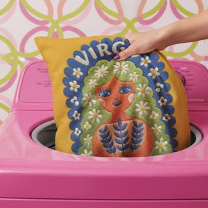 A square throw pillow featuring a vibrant Virgo zodiac design on a golden mustard yellow background. The central illustration shows a terracotta-skinned figure with green hair adorned with white daisies. Blue leaf motifs are printed on the figure’s chest. The word "VIRGO" appears in white bubble letters at the top. The entire design is framed by a royal blue scalloped border, featuring a high-fidelity trompe-l'œil print that mimics 3D embroidery.