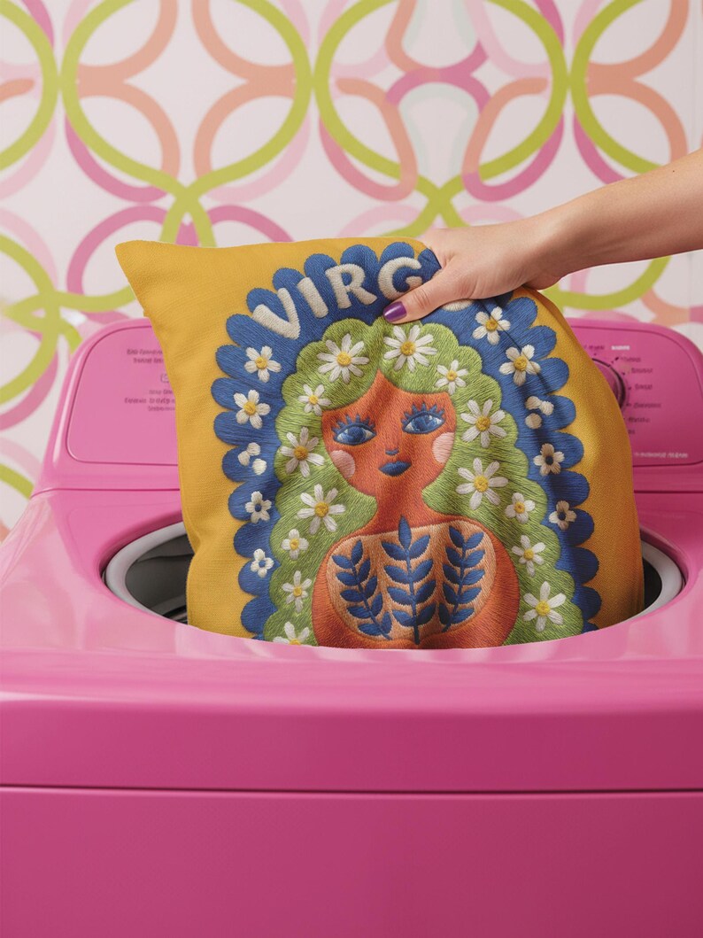 A square throw pillow featuring a vibrant Virgo zodiac design on a golden mustard yellow background. The central illustration shows a terracotta-skinned figure with green hair adorned with white daisies. Blue leaf motifs are printed on the figure’s chest. The word "VIRGO" appears in white bubble letters at the top. The entire design is framed by a royal blue scalloped border, featuring a high-fidelity trompe-l'œil print that mimics 3D embroidery.
