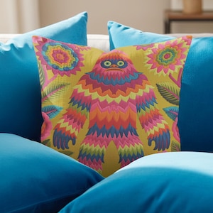 A square faux embroidery pillow featuring a brightly colored maximalist design against a mustard yellow background. In the center stands a stylized, furry Bigfoot or Sasquatch figure rendered in vibrant layers of pink, orange, blue, and lime green with a blue face and yellow eyes. The central creature is surrounded by large, detailed symmetrical pink and blue flowers and leaves. The flat printed fabric creates a highly realistic optical illusion of dimensional crewel embroidery stitches.