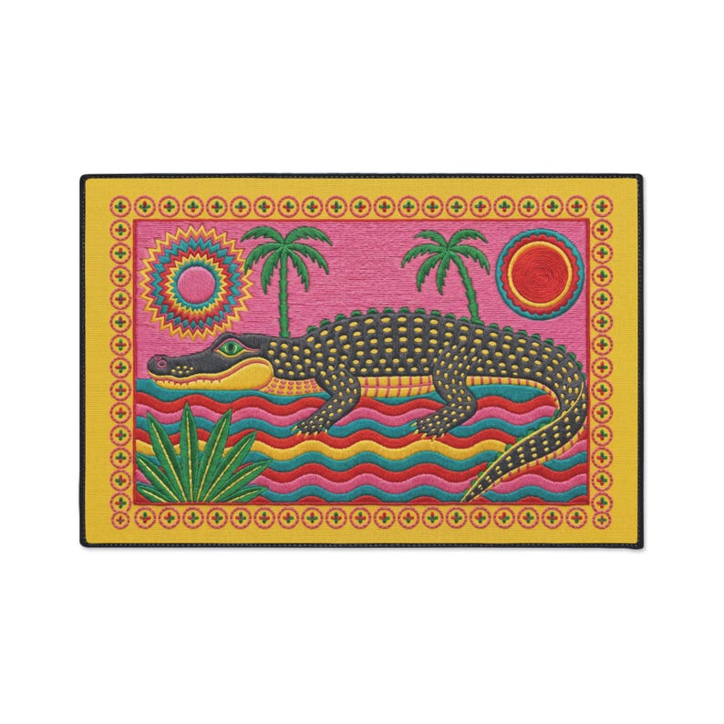 A rectangular doormat featuring a colorful 2D flat print designed to mimic raised, textured yarn embroidery. The central graphic showcases a large dark grey and yellow alligator resting in wavy water stripes of red, bright blue, and yellow. The background is hot pink, decorated with two green palm trees, a leafy green agave plant, and two stylized suns. The entire design is enclosed by a yellow border with repeating circle motifs and finished with a sleek black outer edge border trim.